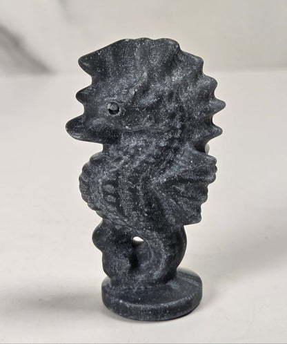 A small dark gray seahorse figurine with a textured, stone-like finish, standing on a round base against a light gray background.