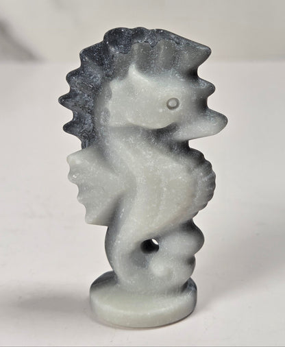 A small carved seahorse figurine in a light gray stone-like material with darker gray shading, standing on a round base against a plain light background.