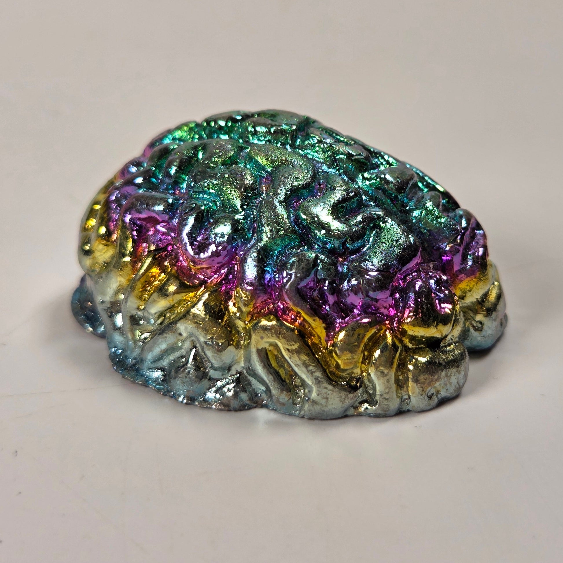 A small, highly textured, brain-shaped object with an iridescent, metallic rainbow finish, resting on a plain light gray surface.