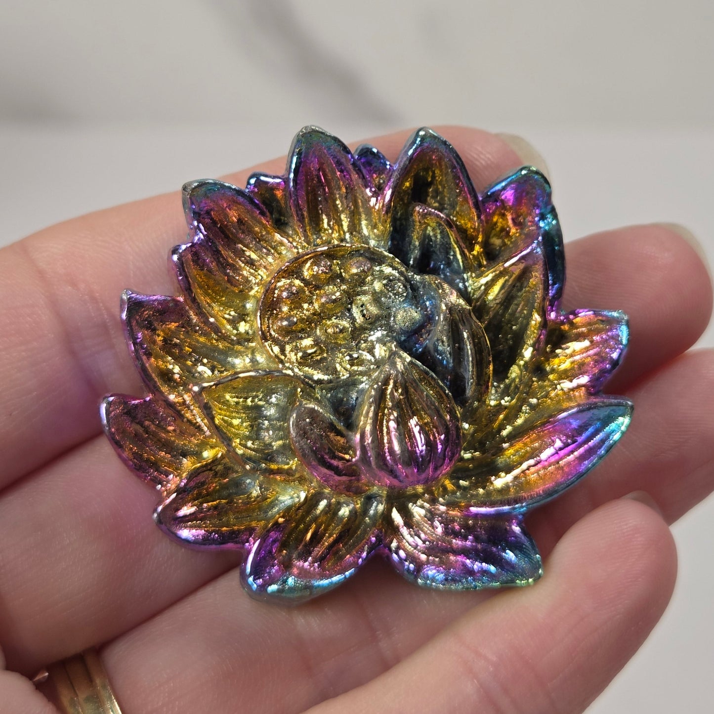 A close-up of a hand holding a small, iridescent lotus flower-shaped accessory, likely a brooch or charm, with a gold-toned center and shifting rainbow metallic petals.