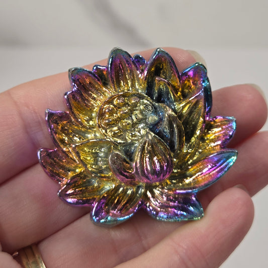 A close-up of a hand holding a small, iridescent lotus flower-shaped accessory, likely a brooch or charm, with a gold-toned center and shifting rainbow metallic petals.