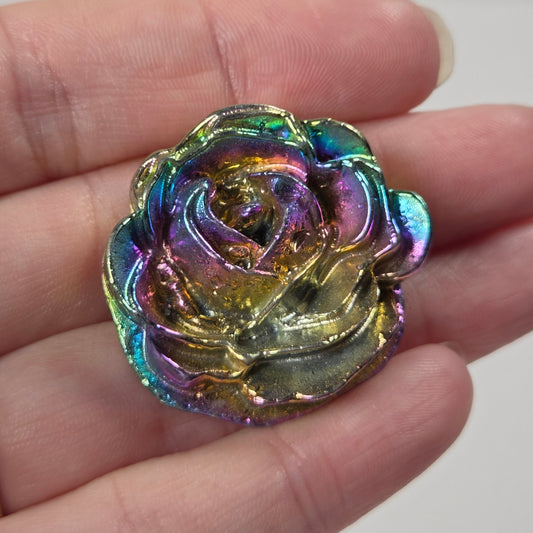 An iridescent rose-shaped decorative piece resting on a person's fingers. The item has a metallic, holographic finish shifting between gold, green, blue, and purple.