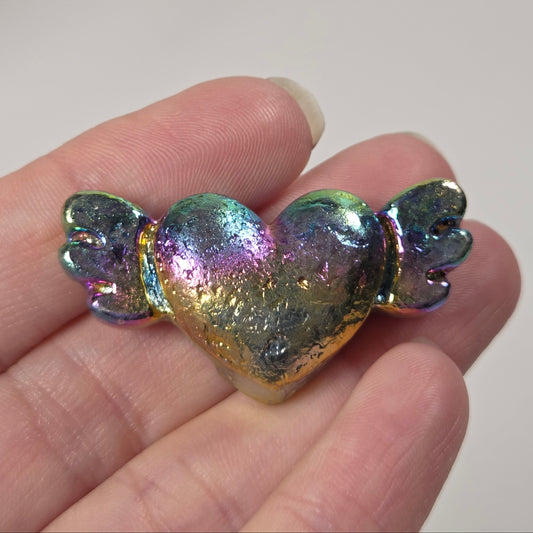 A small iridescent heart-shaped charm with tiny wings, held in a person's hand against a light gray background.