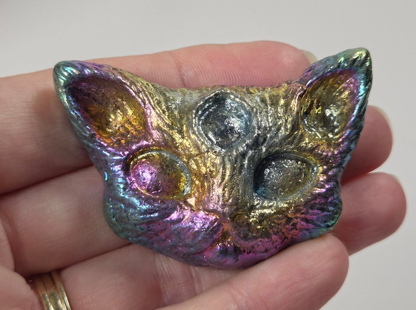 A hand holding a small, cat-shaped metallic pendant with an iridescent, rainbow-like finish that shifts between gold, green, blue, and purple.