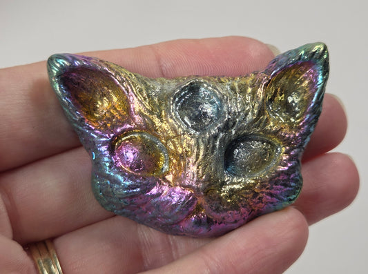A hand holding a small, cat-shaped metallic pendant with an iridescent, rainbow-like finish that shifts between gold, green, blue, and purple.