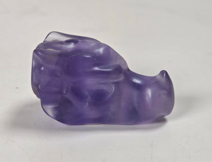 A small carved stone figurine resembling a stylized animal head or creature, made from polished fluorite, standing on a light gray surface.