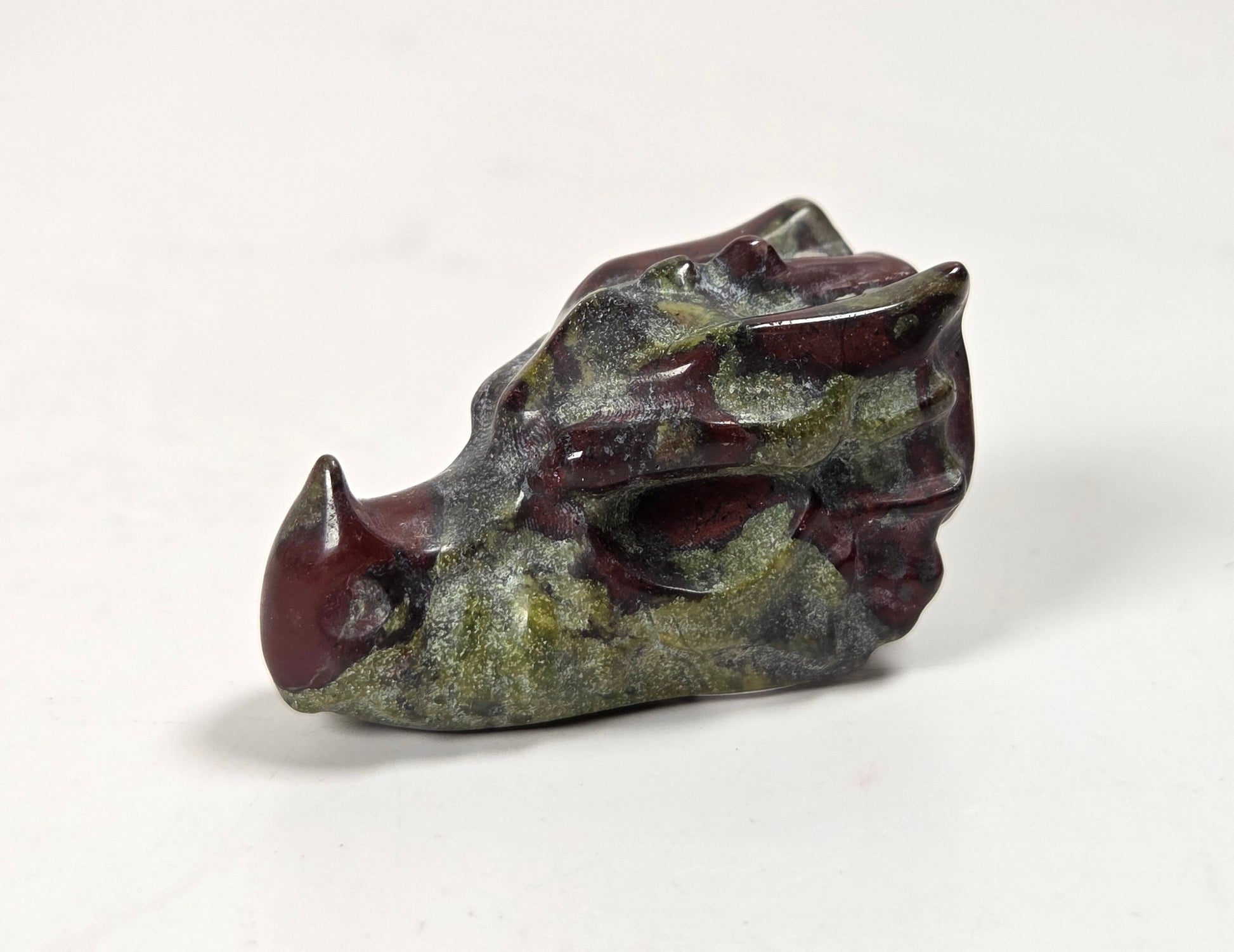 A small carved animal skull figurine, likely a boar or pig, featuring a dark reddish-brown horn and snout with textured green and gray patina details on the head, set against a plain white background.