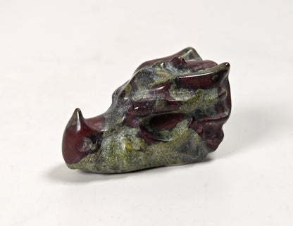 A small carved animal skull figurine, likely a boar or pig, featuring a dark reddish-brown horn and snout with textured green and gray patina details on the head, set against a plain white background.