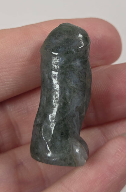 A small carved dark green stone object, shaped like a stylized penis, held in a person's hand against a plain light background.