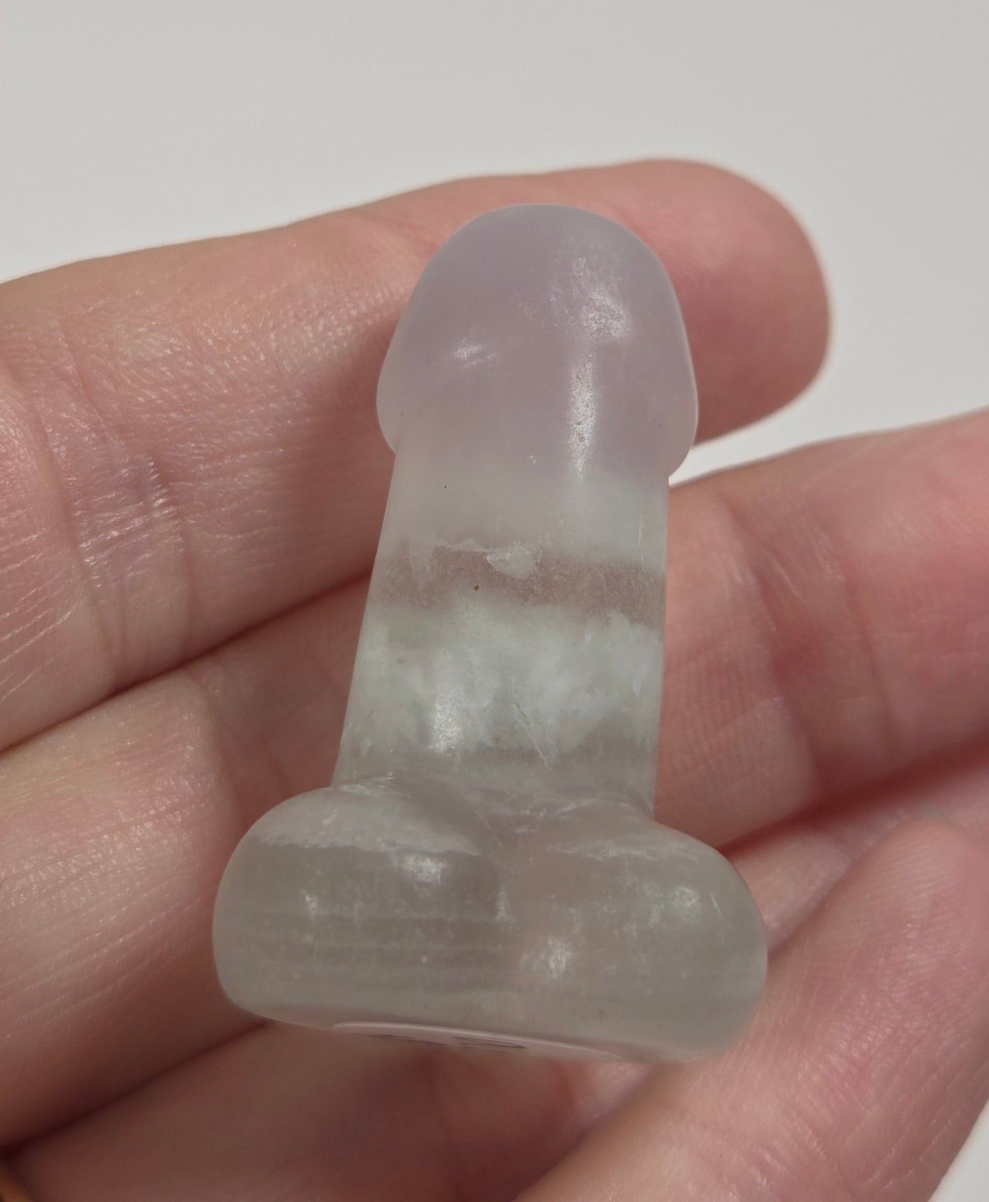 A small translucent pale pink and white stone dildo held in a person's hand against a light gray background.