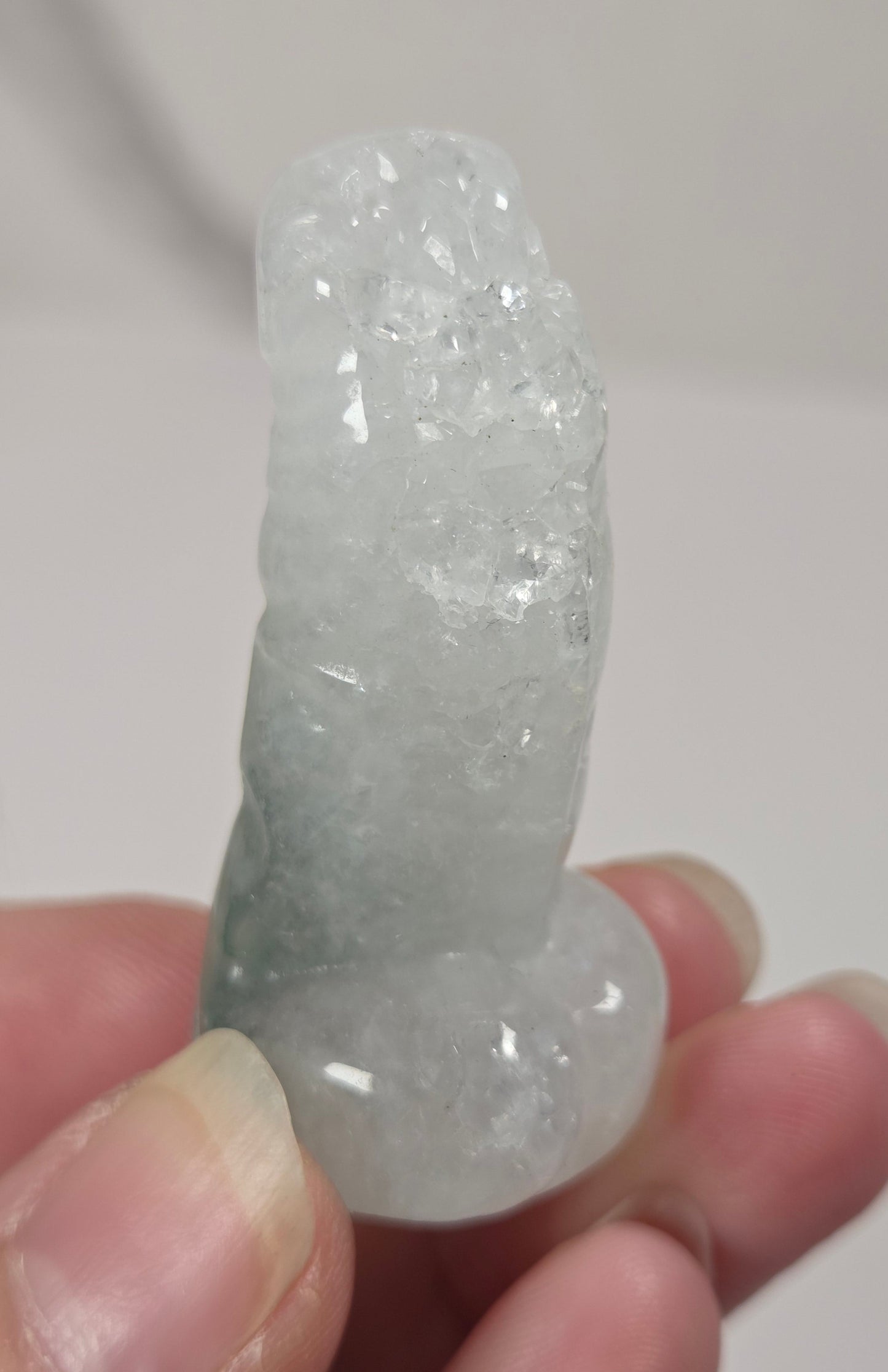 A hand holding a small, rough, pale greenish-white crystal or mineral specimen against a plain light background.