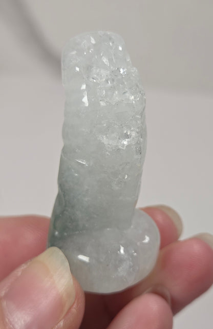 A hand holding a small, rough, pale greenish-white crystal or mineral specimen against a plain light background.