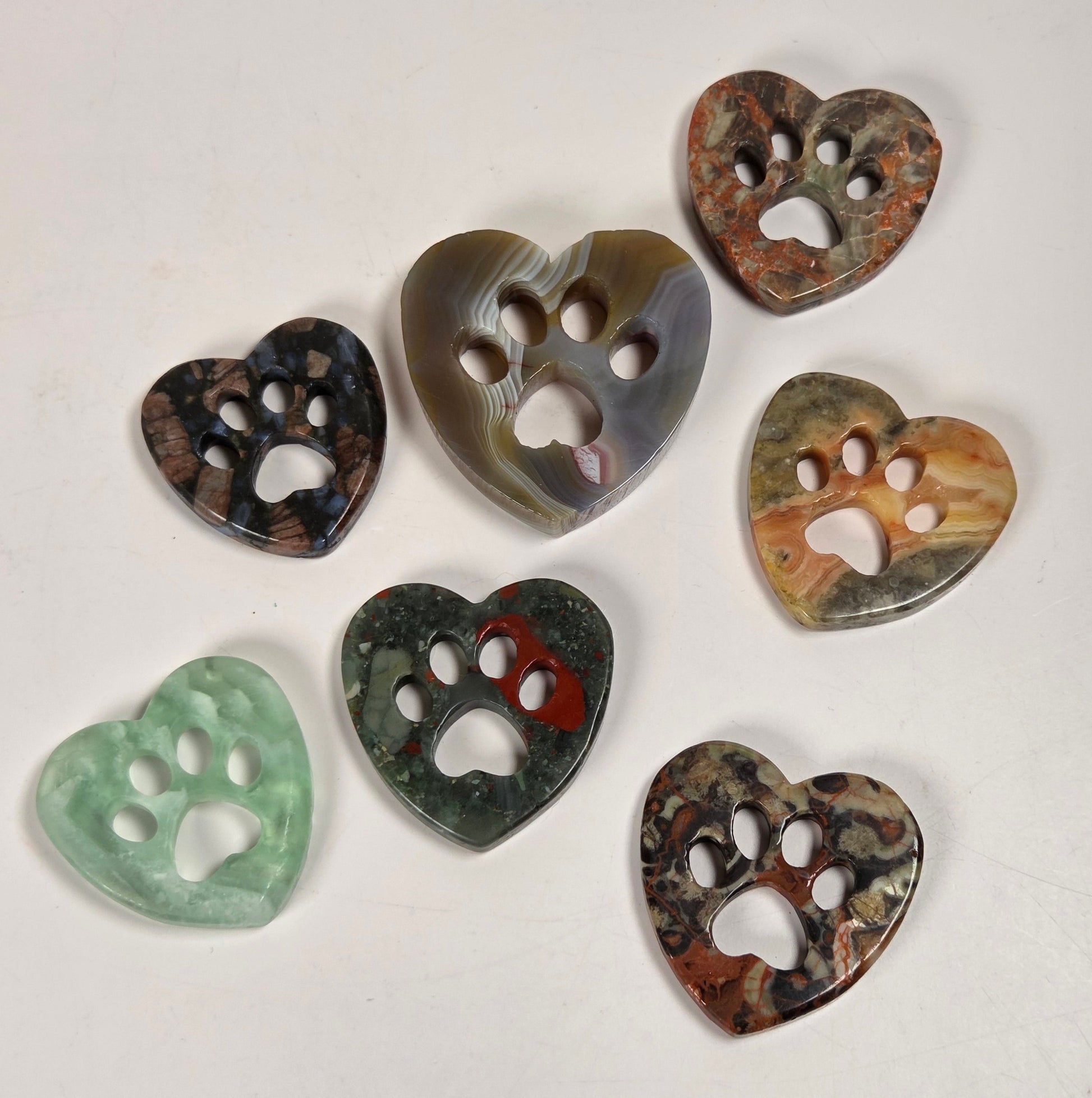 Seven heart-shaped buttons with paw print cutouts, made from various natural stone materials in different colors, arranged on a plain off-white surface.