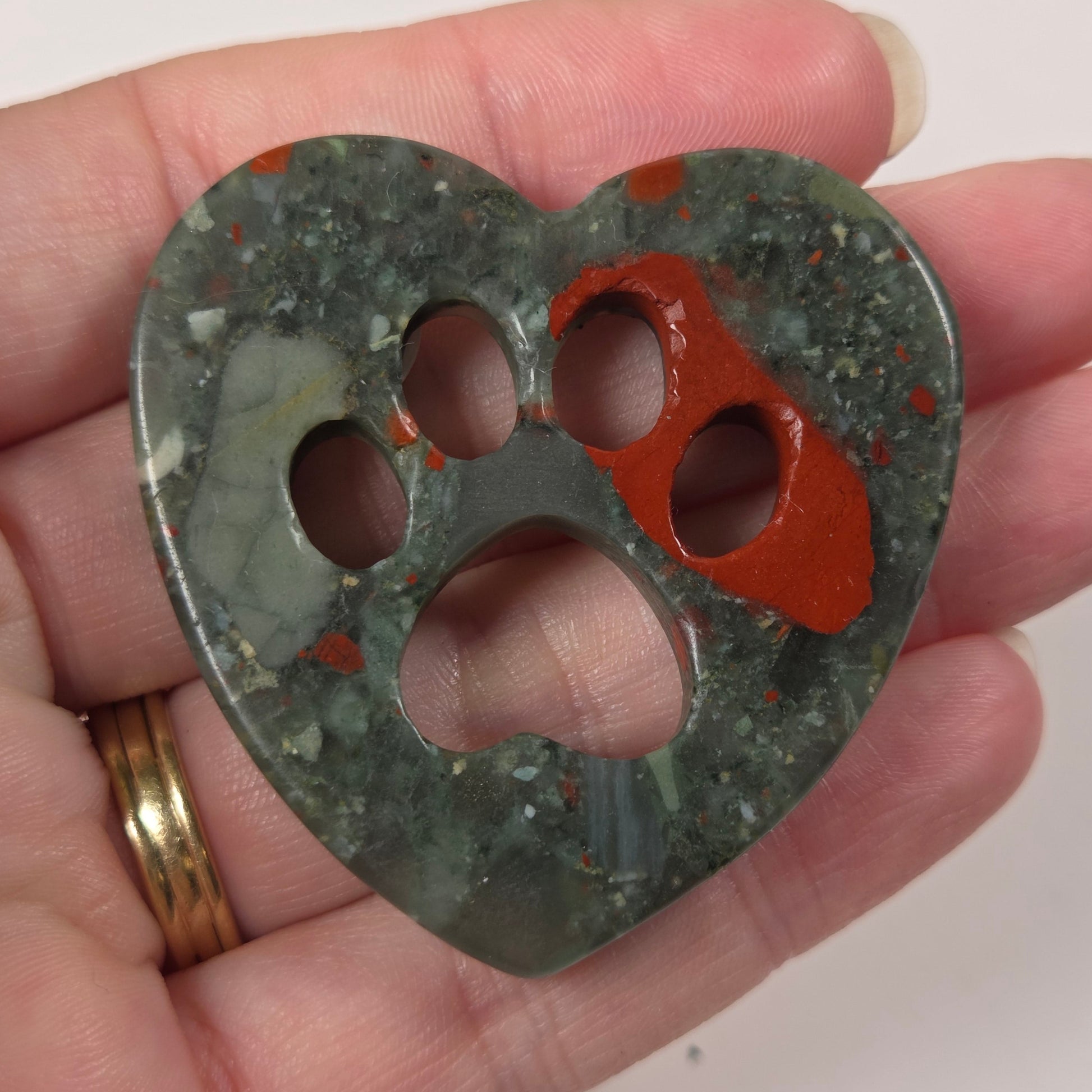 A heart-shaped carved stone pendant with four cutouts, held in a person's hand. The pendant is made of a mottled dark green stone with orange-red inclusions, and the person's hand is visible holding it against a plain light background.