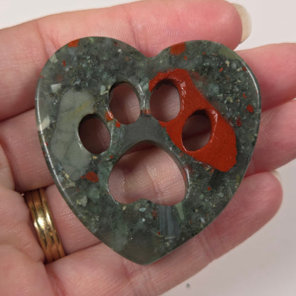 A heart-shaped carved stone pendant with four cutouts, held in a person's hand. The pendant is made of a mottled dark green stone with orange-red inclusions, and the person's hand is visible holding it against a plain light background.