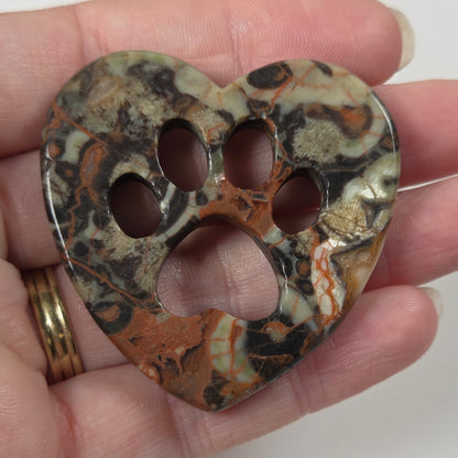 A heart-shaped bone-shaped pendant with a paw print cutout, featuring a marbled pattern of brown, black, beige, and white, held in a person's hand.
