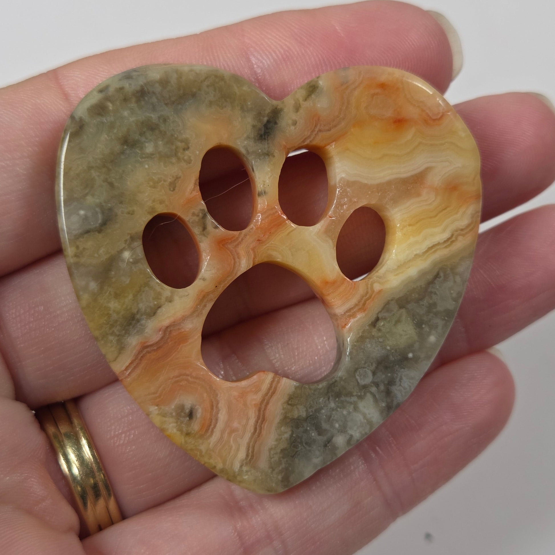 A heart-shaped stone pendant with a paw print cutout, held in a person's hand. The pendant features natural agate-like banding in shades of orange, yellow, green, and brown.