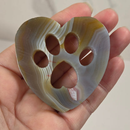 A hand holding a heart-shaped carved stone object with four circular cutouts, showing natural agate-like banding in gray, white, tan, and brown tones.