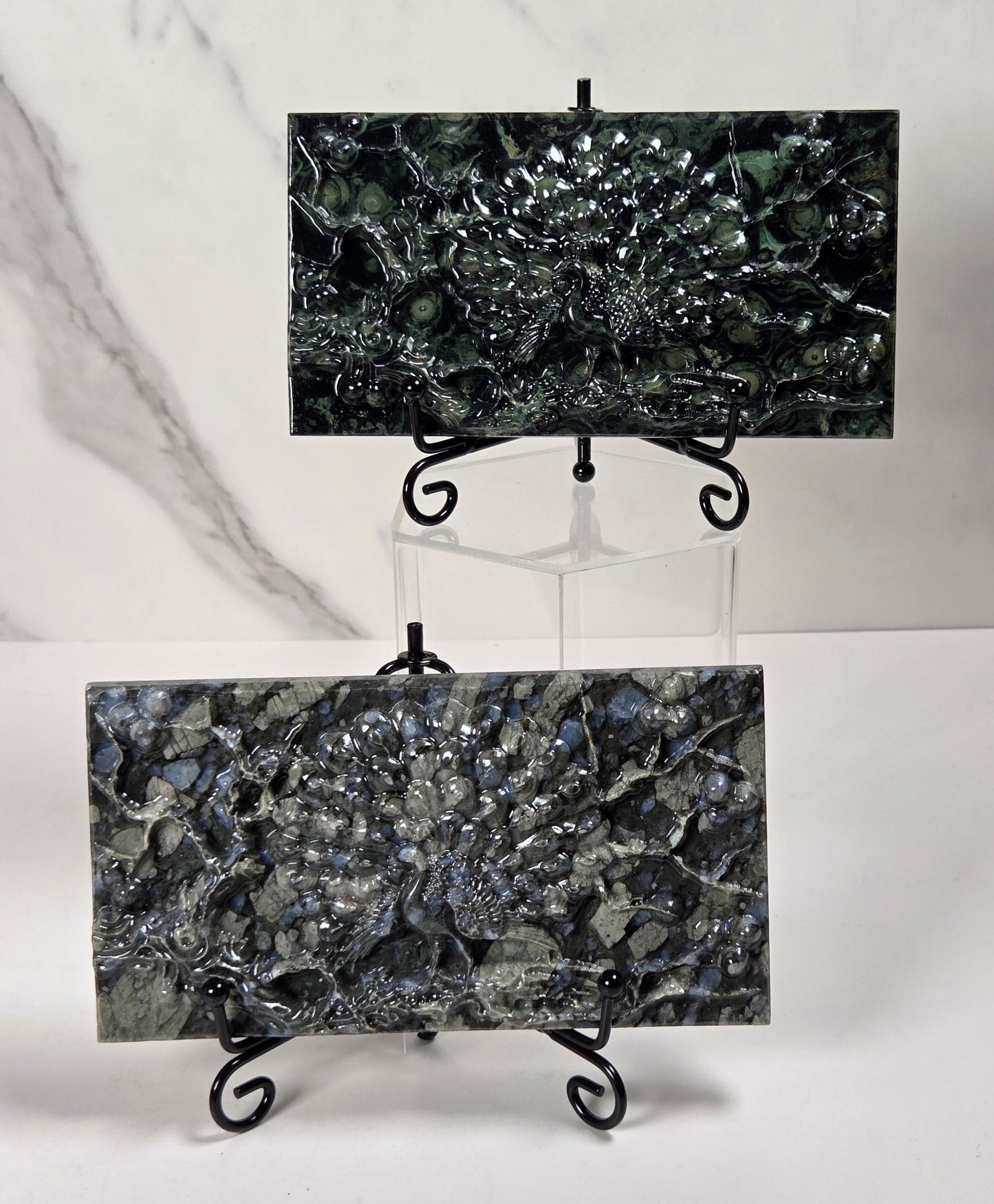 Two rectangular black and gray stone tiles with a metallic, textured pattern, displayed on small black metal easels against a white marble-patterned background.