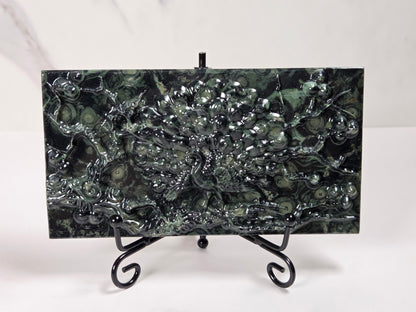 A rectangular dark green malachite stone plaque with a raised peacock design, displayed on a black metal stand against a white marble background.