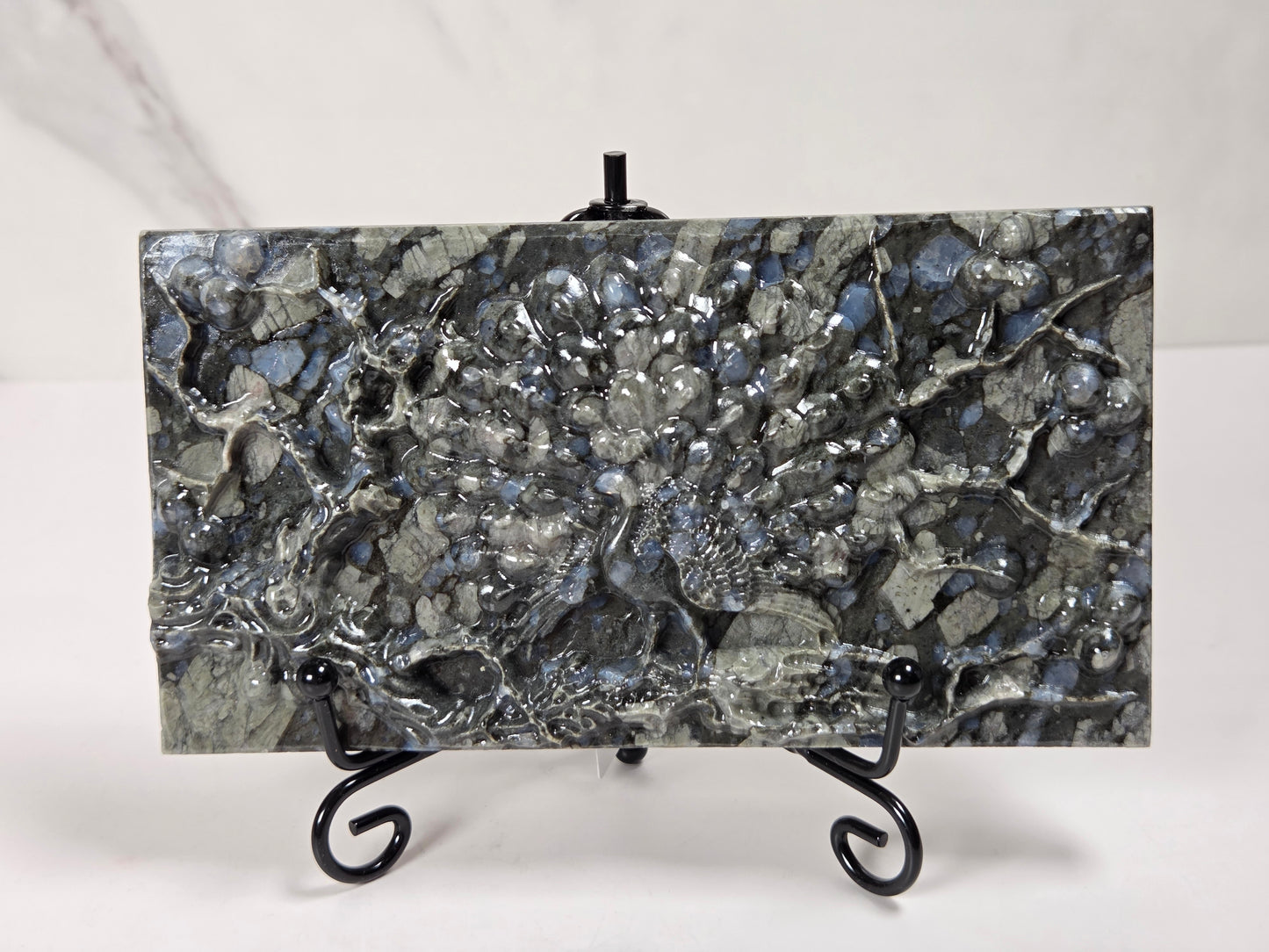 A rectangular polished stone slab with a mottled gray, black, and silver marbled pattern, displayed on a small black metal stand against a light gray background.