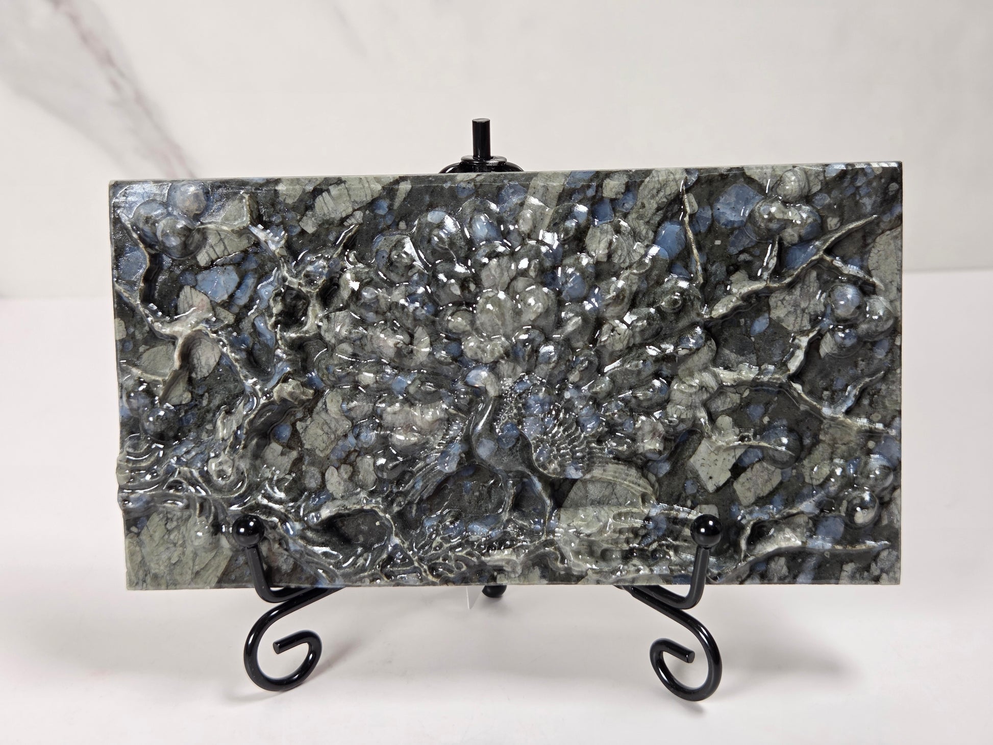 A rectangular polished stone slab with a mottled gray, black, and silver marbled pattern, displayed on a small black metal stand against a light gray background.