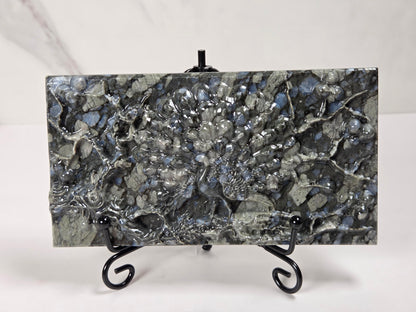 A rectangular polished stone slab with a mottled gray, black, and silver marbled pattern, displayed on a small black metal stand against a light gray background.