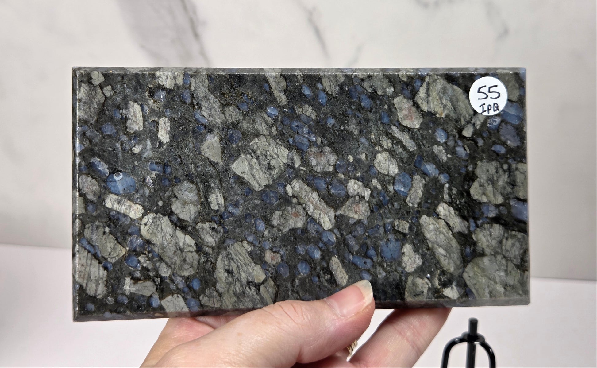 A hand holding a rectangular slab of dark gray and black granite with prominent blue and white flecks, set against a light marble-patterned background.