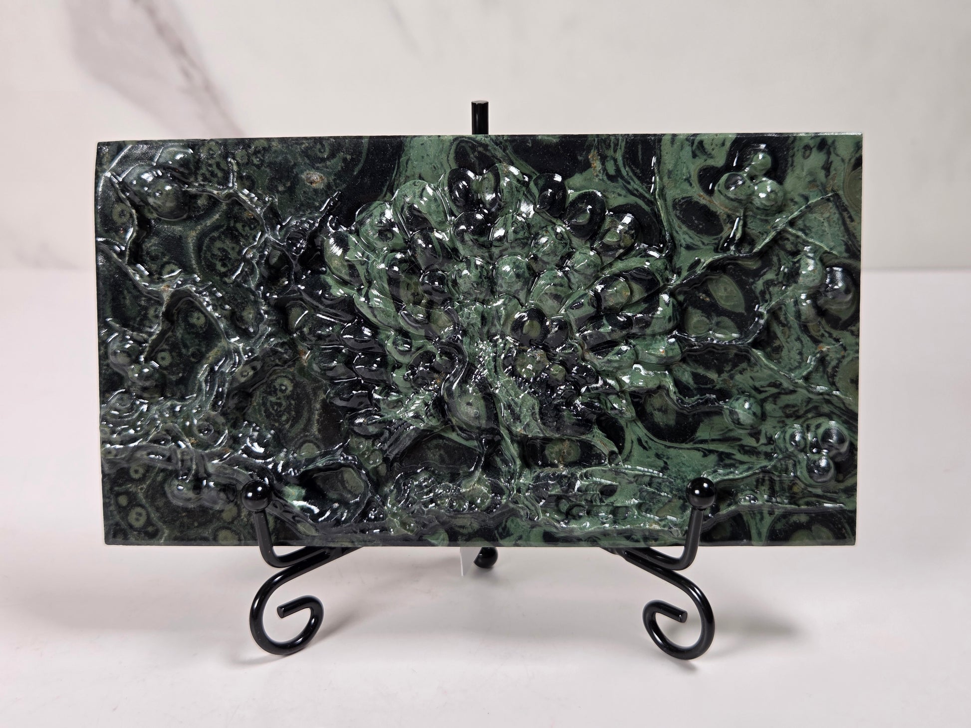 A rectangular green malachite stone wall tile with a raised, textured floral pattern, displayed on a black metal stand against a white background.