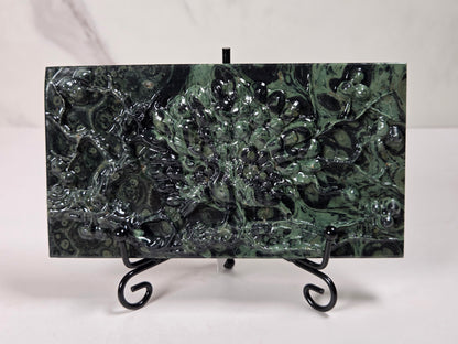 A rectangular green malachite stone wall tile with a raised, textured floral pattern, displayed on a black metal stand against a white background.