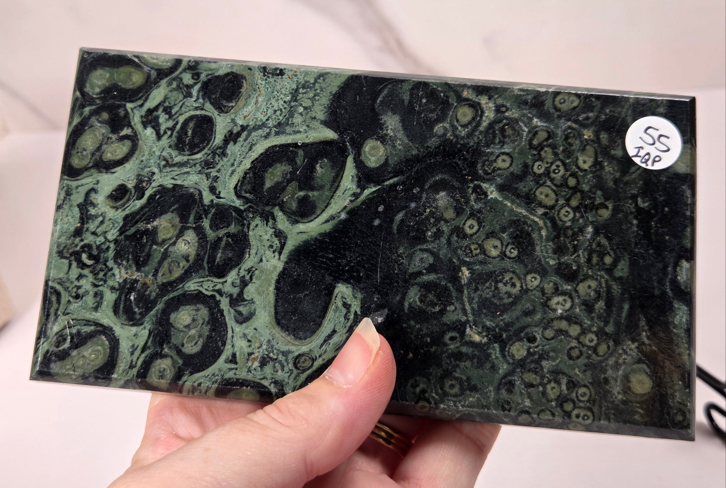 A hand holding a rectangular polished slab of dark green and black mottled stone or marble with organic veining patterns. A small white sticker reading 'SS 148' is visible on the top right of the slab.