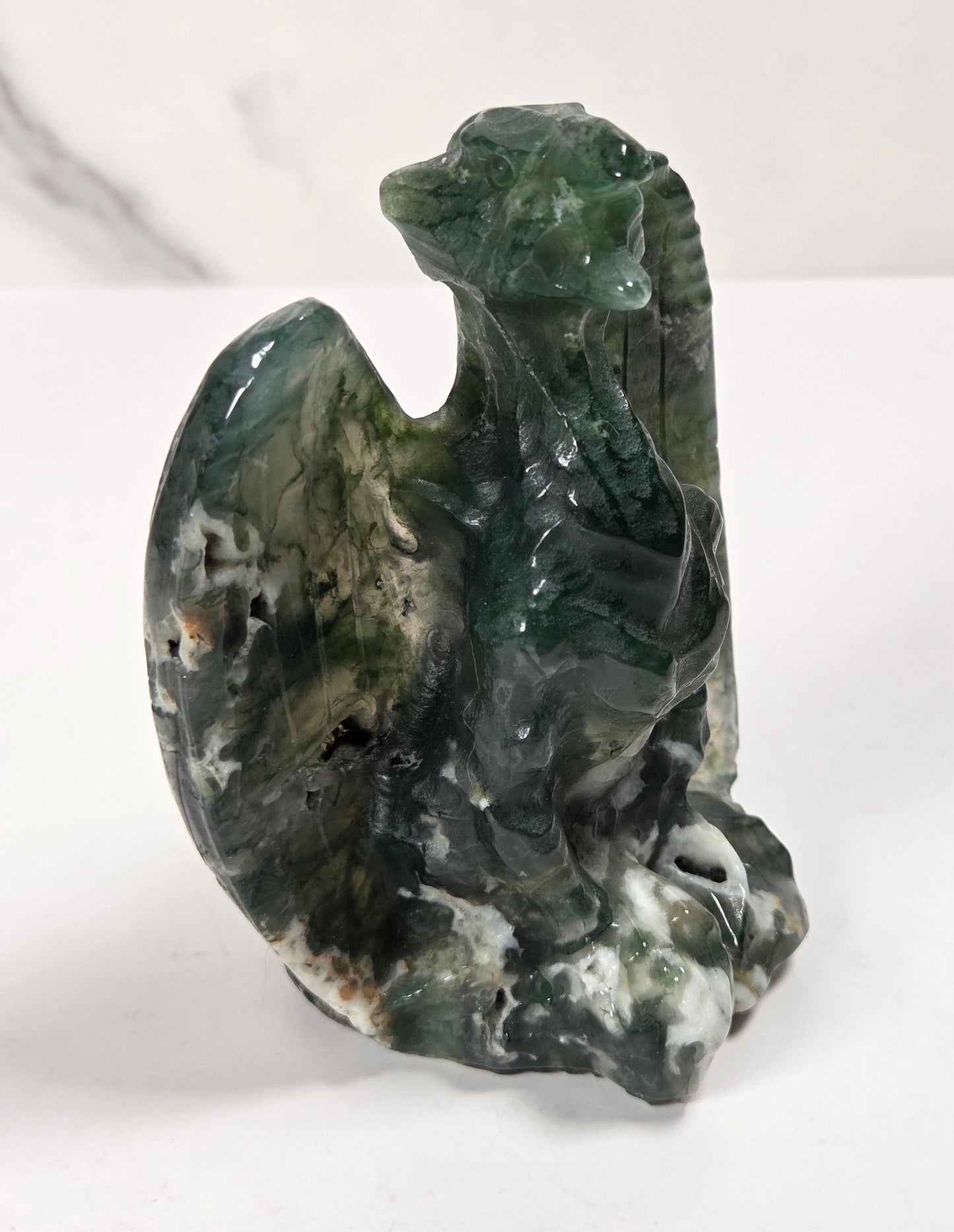 A carved green stone dragon figurine with natural marbling, standing on a white surface against a light gray marble-patterned background.
