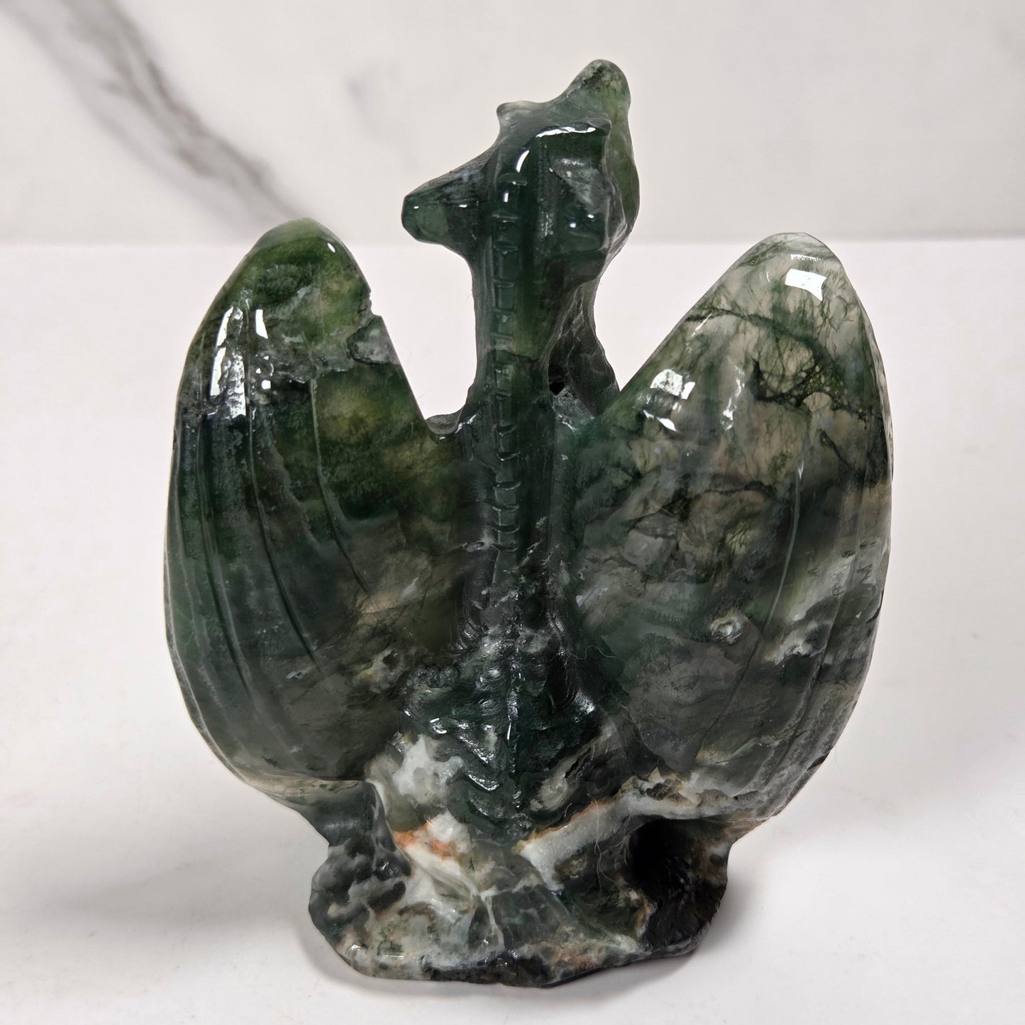 A polished dark green stone carving shaped like a stylized bird or phoenix with wings spread, photographed on a light surface against a pale marble-like background.