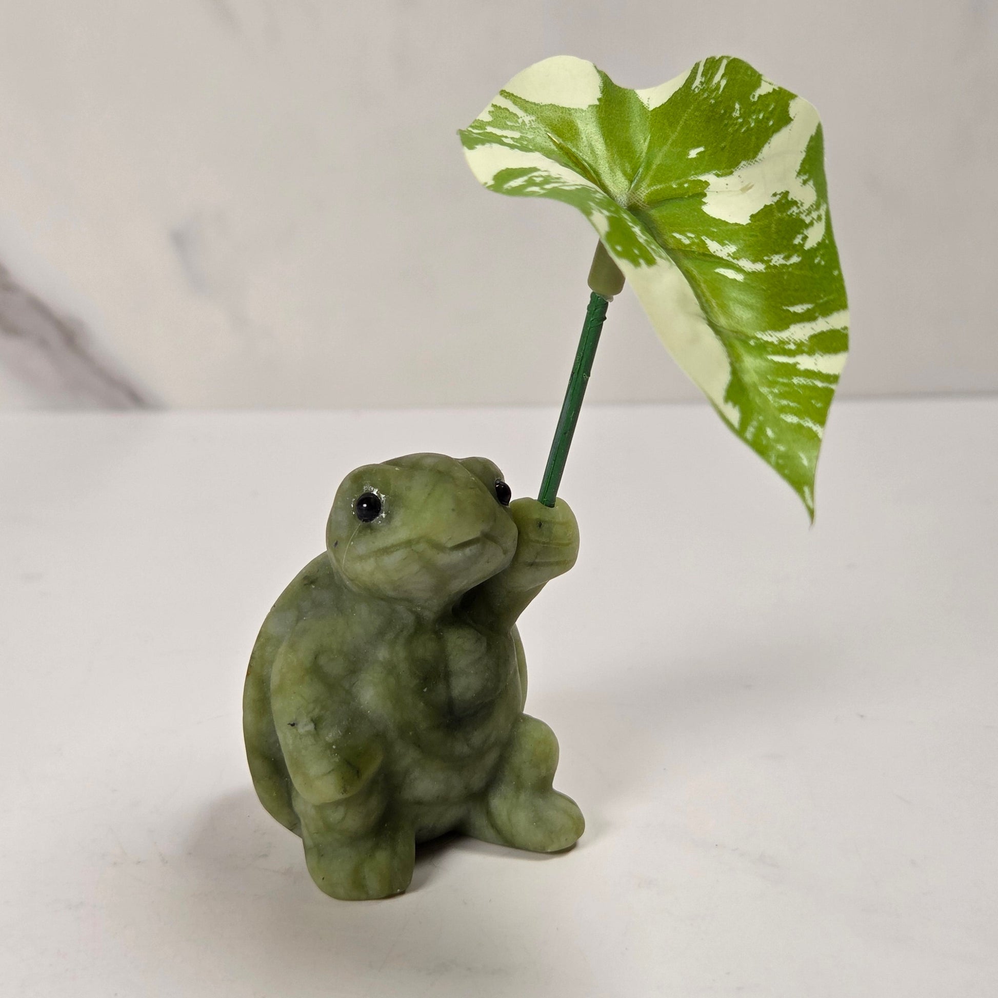 A small green turtle figurine holding a variegated green and cream plant leaf on a stem, set against a light gray marble-patterned background.