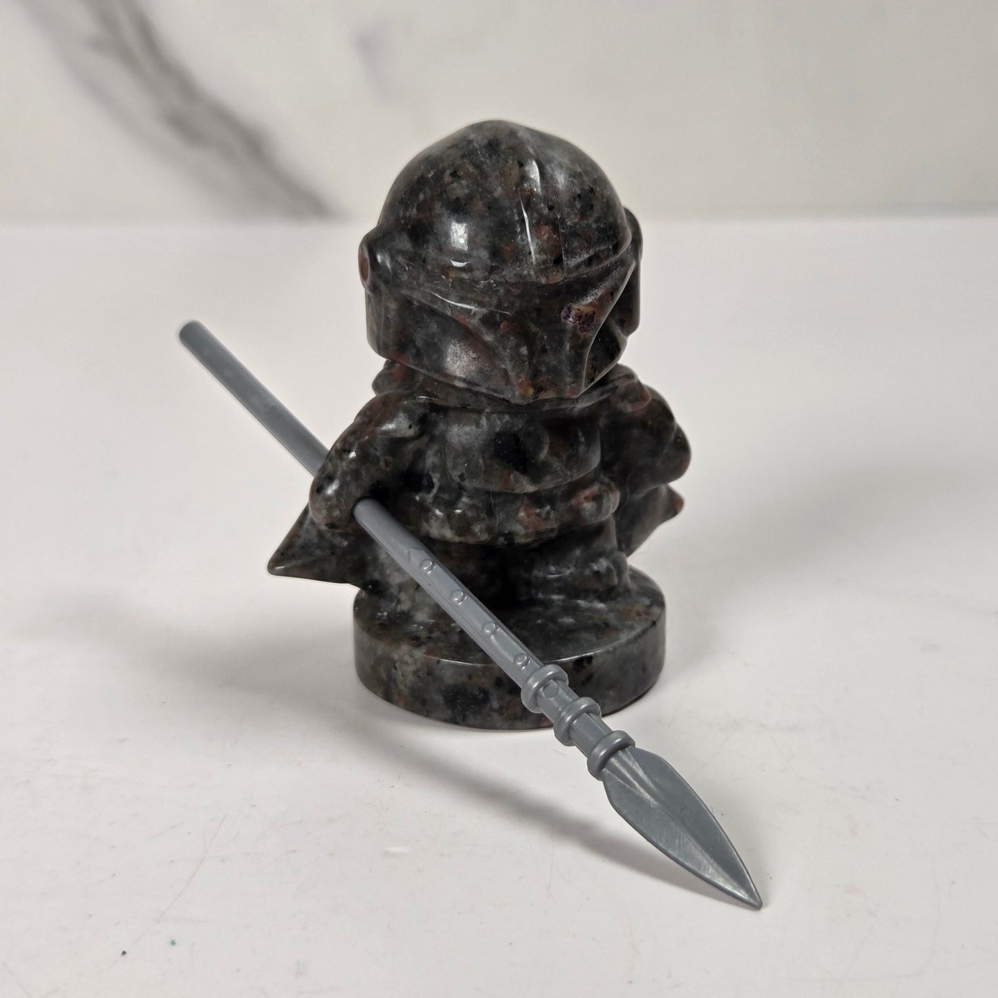 A small black and dark gray stone figurine of a helmeted character holding a gray spear, photographed on a light gray surface with a marble-patterned background.