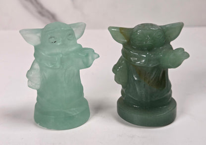 Two small green character figurines, one translucent pale mint green and one opaque dark green, displayed side by side on a white surface against a light gray marble-patterned background.