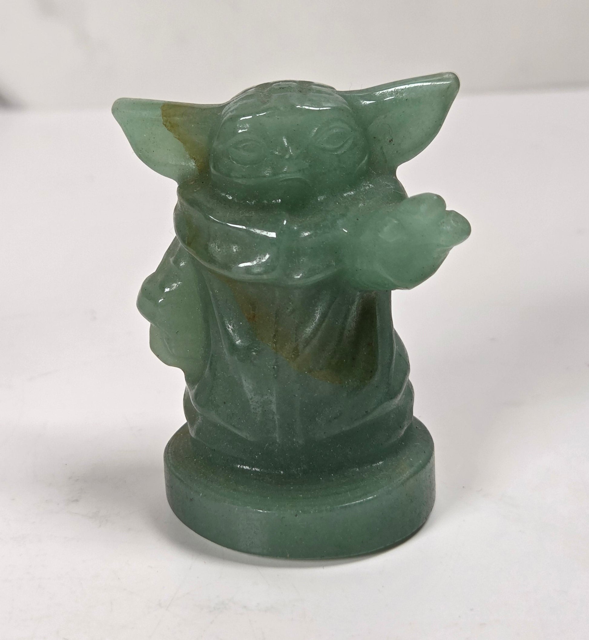 A small green stone figurine of a child-like Jedi or Yoda-like character, standing on a round base against a white marble-patterned background.