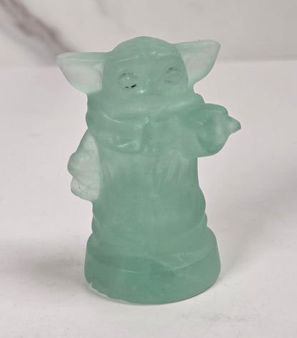 A small translucent mint green figurine of Grogu, standing on a white surface against a light gray and white marble-patterned background.