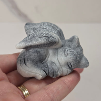 A small gray stone figurine of a sleeping fox or rabbit with wings, held in a person's hand. The figurine has a textured, matte stone finish with gray and white marbling.