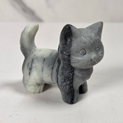 A small carved cat figurine made from split stone, with one half in light gray and the other half in dark charcoal gray, standing on a white surface.