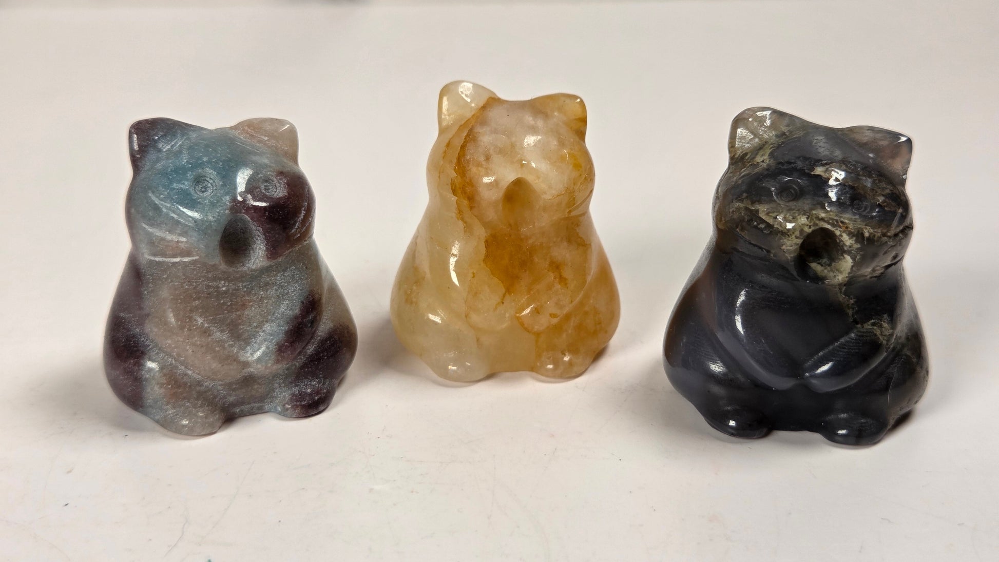 Three small carved stone figurines shaped like caTS, displayed side by side on a light surface. The left bear is gray with blue and purple marbling, the center bear is translucent amber-brown, and the right bear is dark black with gray veining.
