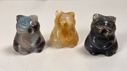 Three small carved stone figurines shaped like caTS, displayed side by side on a light surface. The left bear is gray with blue and purple marbling, the center bear is translucent amber-brown, and the right bear is dark black with gray veining.