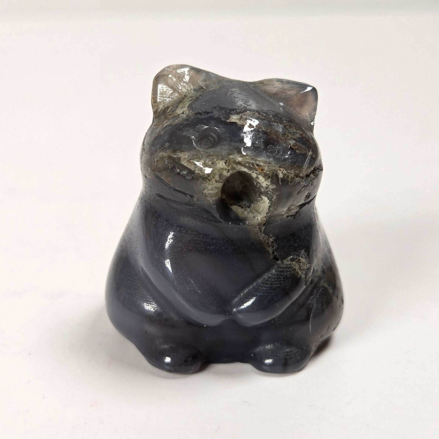 A small carved figurine of a cat in a sitting position, made from polished dark gray stone with natural lighter gray and beige inclusions, set against a plain off-white background.