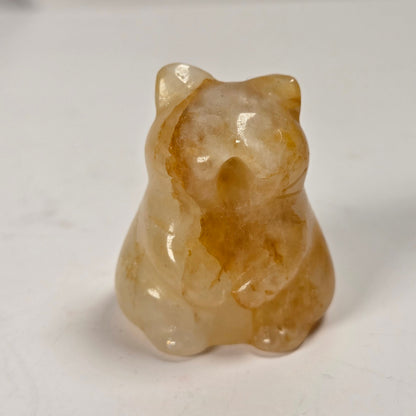 A small carved figurine of a cat made from polished translucent stone, showing warm amber and cream tones, set against a plain light background.