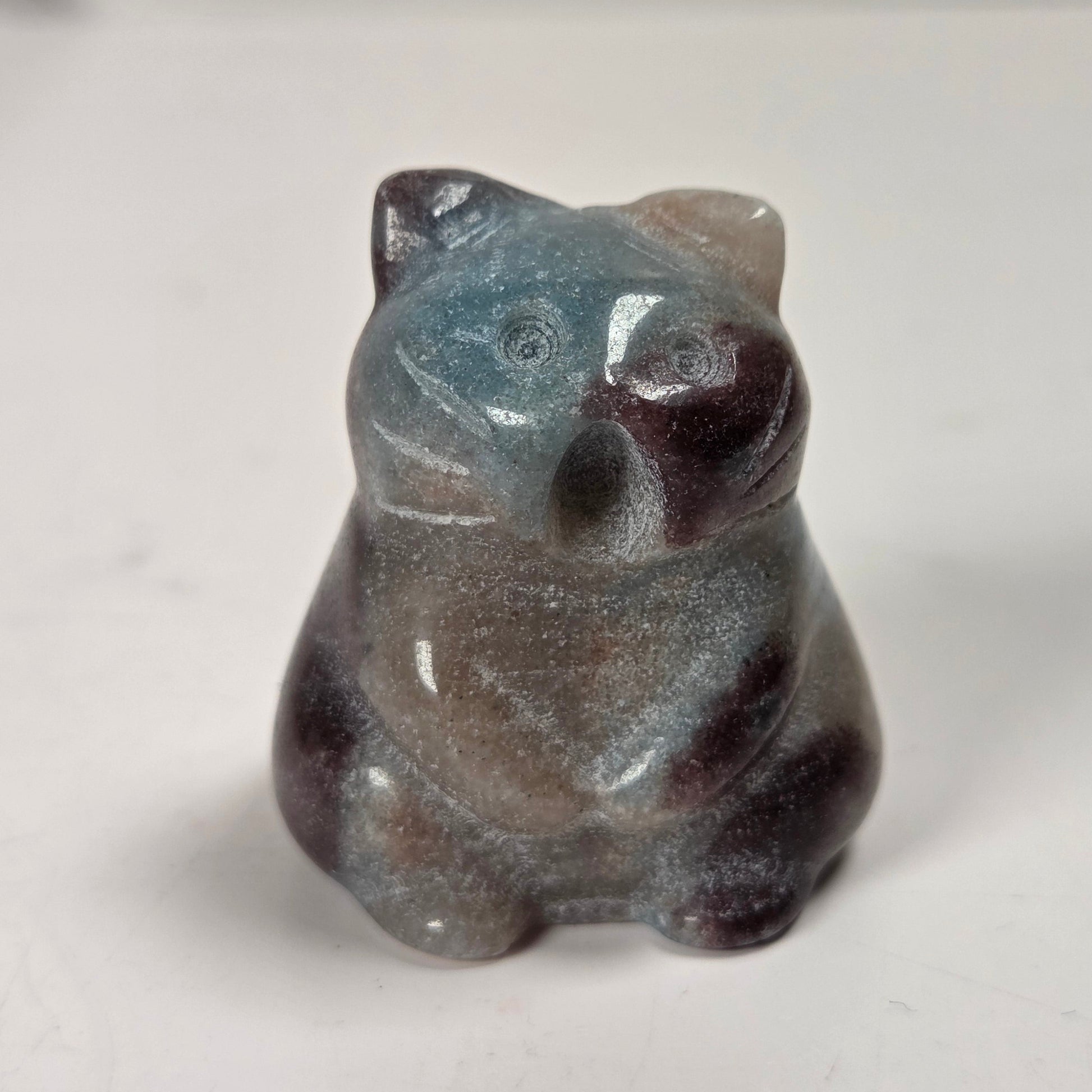 A small carved figurine of a cat made from a speckled stone with blue, gray, brown, and dark reddish tones, sitting on a plain light surface.