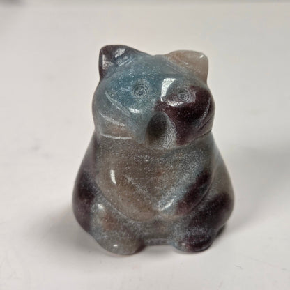 A small carved figurine of a cat made from a speckled stone with blue, gray, brown, and dark reddish tones, sitting on a plain light surface.