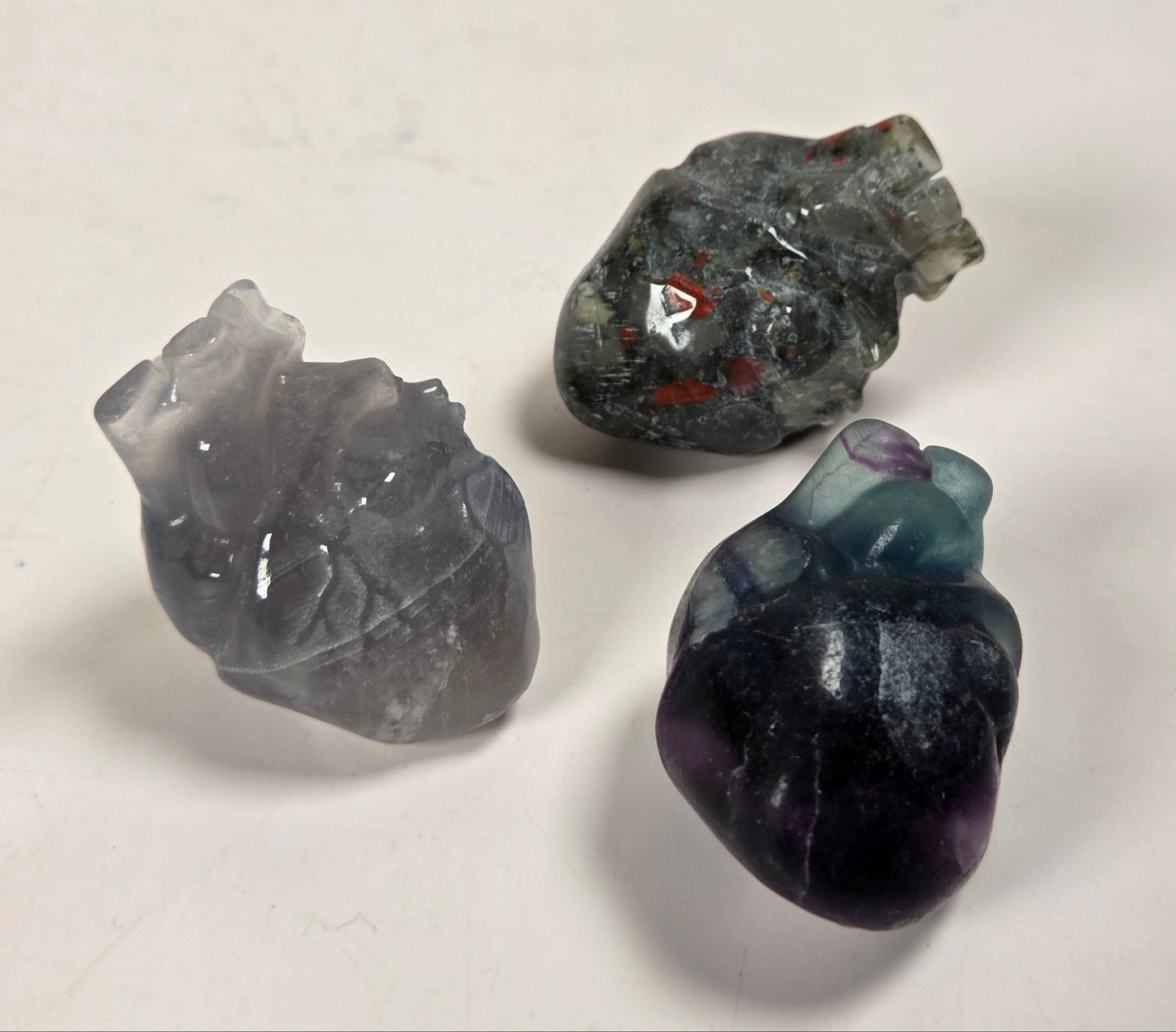 Three small polished stone heart-shaped figurines in gray, dark gray with red inclusions, and dark purple with blue-green highlights, arranged on a plain off-white surface.