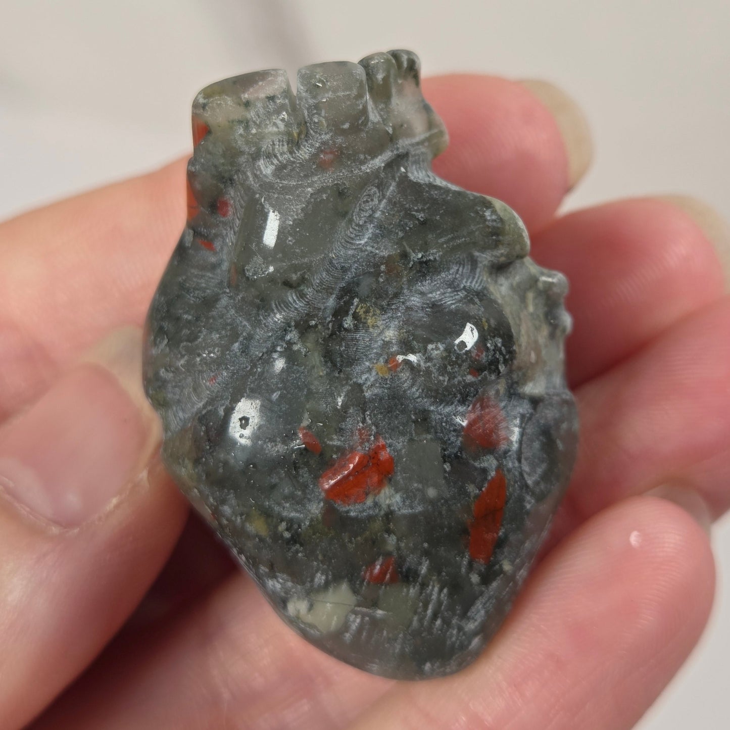 A hand holding a small carved translucent heart-shaped stone pendant with red inclusions, photographed against a softly blurred light background.