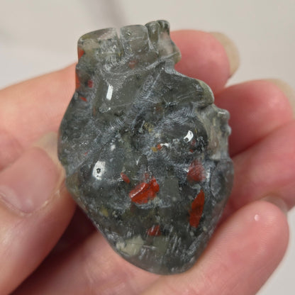 A hand holding a small carved translucent heart-shaped stone pendant with red inclusions, photographed against a softly blurred light background.