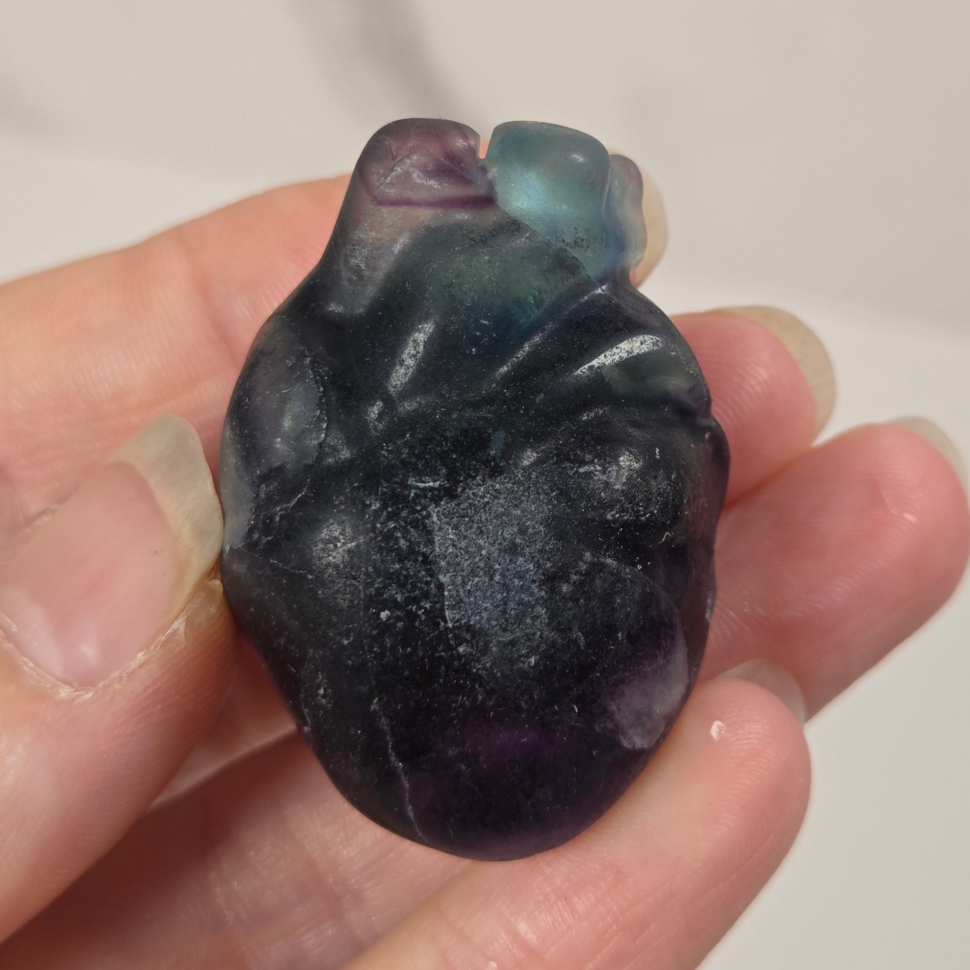 A hand holding a small polished heart-shaped stone with a dark purple and black body and blue-green highlights near the top, against a softly blurred light background.